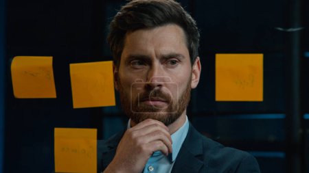 Foto de Thoughtful Caucasian businessman employee middle-aged man analyze information on stickers read sticky notes on glass wall standing in office planning project manage tasks startup ideas organizes work - Imagen libre de derechos