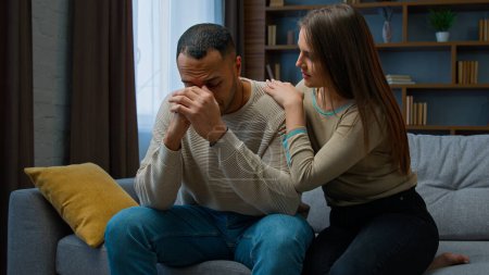 Photo for Desperate sad man sit on sofa upset with bad news complain life problem caring wife support boyfriend. Apologizing woman say sorry hugging annoyed husband diverse couple talking after quarrel conflict - Royalty Free Image