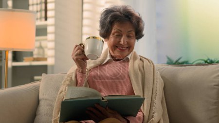 Téléchargez les photos : Happy souriant inspiré Caucasienne vieille femme couverte de couverture sur canapé boire du thé café avec tasse livre de lecture sourire atmosphère paix dame âgée retraitée lire boisson à la maison canapé - en image libre de droit