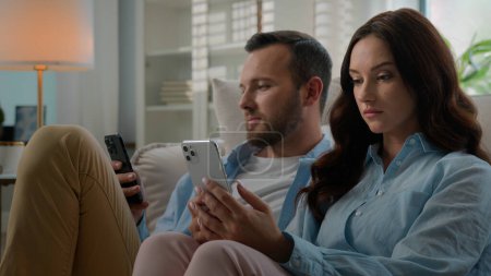 Foto de Involved Caucasian couple man and woman at home using phones mobile app reading devices social media addicted people at living room browsing smartphones internet addiction family interested in gadgets - Imagen libre de derechos