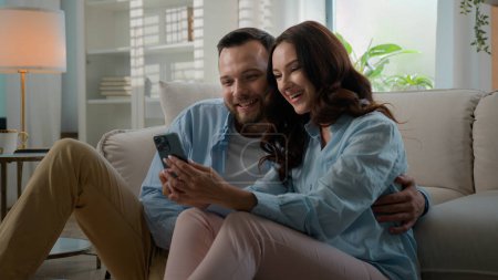 Téléchargez les photos : Millennial couple Caucasian man woman married husband wife at home talking using smartphone together happy family discuss offers choosing shopping online with mobile phone boyfriend showing thumb up - en image libre de droit