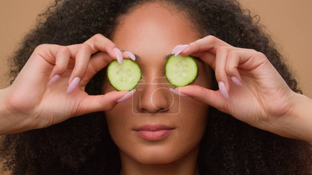Photo for Portrait happy young beautiful African American woman girl with fresh cucumber slices eyes cover smiling laughing perfect skin care moisturizing refresh on beige studio background organic cosmetics - Royalty Free Image