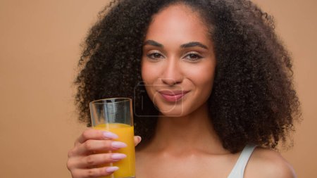 Téléchargez les photos : Heureuse femme afro-américaine en bonne santé modèle féminin boire du jus d'orange frais boisson vitaminée étirer la main avec du verre motiver cliquetis suggérer d'essayer régime désintoxication soins de santé en studio - en image libre de droit