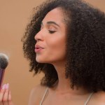 Close up portrait African American ethnic woman model girl holding brush blow away dust apply skin tone powder to nose smiling at camera at beige studio background beautiful lady cosmetics make-up