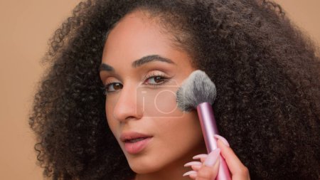 Photo for Close up portrait at beige studio background beautiful African American woman attractive ethnic girl looking at camera apply powder for face make-up preparing cosmetics beauty blush bronzer contouring - Royalty Free Image