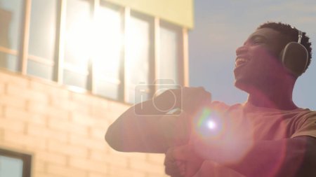 Foto de Feliz baile de bailarín masculino en el cielo soleado en la ciudad danza despreocupada hombre afroamericano diversión al aire libre movimiento para cantar escuchando música auriculares chico en auriculares riendo manos móviles en las luces del sol - Imagen libre de derechos