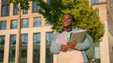 Photo for African American woman university college academy campus student teen girl high school schoolgirl with books schoolbag headphones walking in city outdoors smiling after education studying lesson class - Royalty Free Image