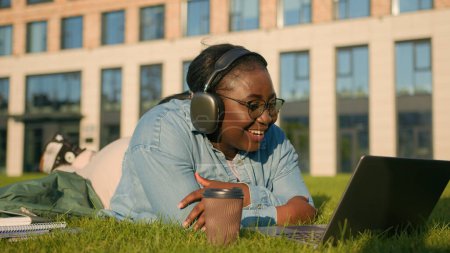 Photo for African American happy joyful woman university college academy student teen girl high school in headphones lying on green grass in park talking video chat call laptop computer conference chat laughing - Royalty Free Image