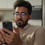 Arabian Indian ethnic man guy use mobile phone on couch browsing mobile internet at home kitchen take break confused read bad news on smartphone male freelancer scrolling cellphone app gadget addict
