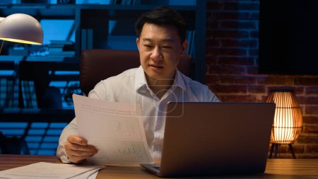 Téléchargez les photos : Focalisé homme d'affaires coréen d'âge moyen travaillant projet en ligne souriant travailleur asiatique à l'aide de logiciels d'entreprise ordinateur portable travail paperasse vérifier les documents au bureau bureau le soir - en image libre de droit