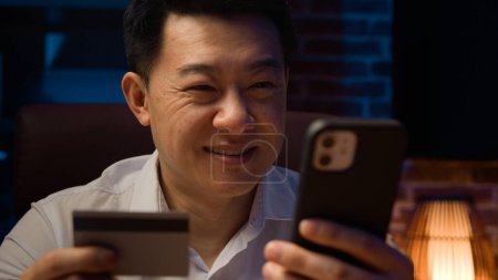 Foto de Sonriente hombre asiático feliz en la noche oscura oficina en casa con teléfono celular utilizando la tarjeta de crédito coreano hombre de negocios hacer orden de reserva de pago sin preocupaciones con fácil descuento de compras de teléfonos móviles en línea - Imagen libre de derechos