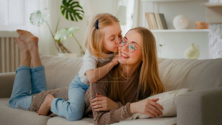 Photo for Happy motherhood family bonding affectionate love Caucasian mother lying on couch sofa daughter little girl child piggyback kiss mom kissing lovely parent warm communication talking sweet tenderness - Royalty Free Image