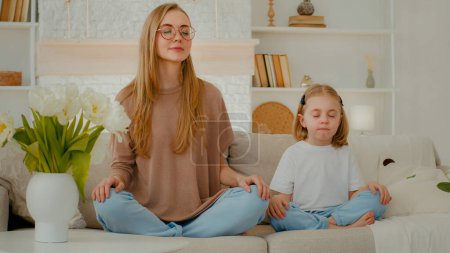 Téléchargez les photos : Caucasian mother with little daughter sitting at home on couch in lotus pose meditate talking. Mom woman in eyeglasses teach preschool child girl meditation sit with closed eyes meditating together - en image libre de droit