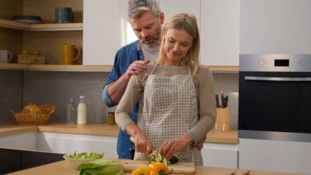 Photo for Happy family Caucasian middle-aged adult at kitchen woman cook vegetable salad cut cucumber husband man hugging cuddling embracing wife couple talking discuss diet food affectionate love talk cooking - Royalty Free Image