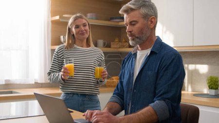 Photo for Caucasian adult woman pour orange juice give glass husband man working online with laptop in home kitchen clinking glasses of fresh talking drinking healthy drink. Middle-aged mature family couple - Royalty Free Image