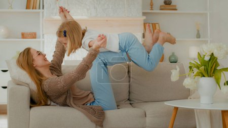 Téléchargez les photos : Happy Caucasian mommy playing with cute adorable toddler child daughter mother holding lifting funny little kid girl up having fun fly at mum knees gymnastics exercise airplane game on sofa at home - en image libre de droit