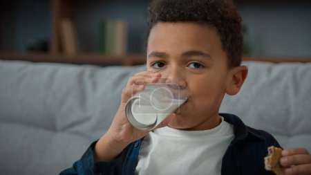 Photo for Close up portrait little funny cute kid boy ethnic child drink milk with cookies at home looking to camera. African American son drinking glass of sweet organic yogurt eating biscuit breakfast eat - Royalty Free Image