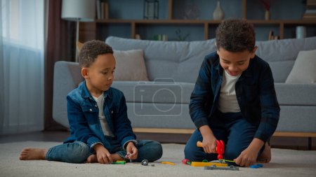Photo for Two African American brothers little ethnic boys children siblings friends kids at home floor in living room playing with toy tools repairing building with hammer having fun busy play game with toys - Royalty Free Image