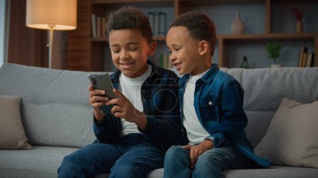 Photo for Children gadget addiction two happy laughing African American little boys brothers browsing mobile phone watching cartoons online without parental control at home couch kids siblings using smartphone - Royalty Free Image