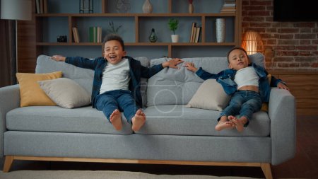 Foto de Happy African American ethnic little boys children carefree active funny kids jumping on soft sofa laugh enjoy relax jump rest on cozy couch moving day to own new home playing game homeowners family - Imagen libre de derechos
