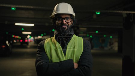 Photo for Indian man handyman worker contractor hands crossed posing in parking smiling mechanic in helmet, glasses power pose at factory warehouse manufacture. Arabian male builder engineer fitter in hard hat - Royalty Free Image