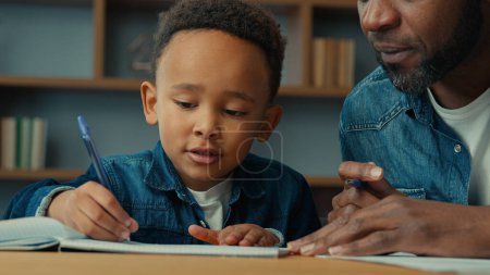 Foto de Afro-americano niño lindo niño colegial con padre adulto étnico escribiendo tarea. Papá ayuda hijo con lección clase escribir explicar ejercicio tarea niño cerrar oídos educación primaria - Imagen libre de derechos