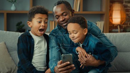 Téléchargez les photos : Afro-Américain père homme avec des petits garçons enfants assis ensemble sur canapé en utilisant smartphone téléphone mobile regarder la vidéo en ligne papa avec préscolaire émotionnel surpris enfants regardent téléphone portable - en image libre de droit
