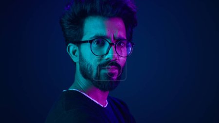 Photo for Indian man guy profile turning head looking camera serious developer coding internet technology hacker programmer Arabian male in glasses neon blue ultraviolet studio background high-tech cyberspace - Royalty Free Image