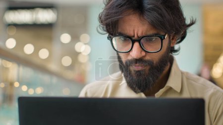 Photo for Serious Indian man in glasses looking at laptop screen working online indoors. Close up Arabian businessman in eyeglasses chatting with computer executive analyze business financial data with Internet - Royalty Free Image