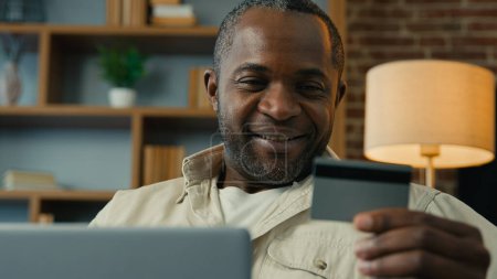 Téléchargez les photos : Heureux homme afro-américain tenant carte de crédit en utilisant le paiement instantané facile sur ordinateur portable à la maison à distance faire des achats dans la boutique en ligne souriant client masculin payer pour le service payant application e-banking - en image libre de droit