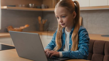 Photo for Caucasian little school girl child baby kid schoolgirl daughter at home kitchen using laptop browsing chatting study internet lesson class studying learning distant with computer playing online game - Royalty Free Image