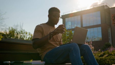 Téléchargez les photos : En plein air étudiant homme afro-américain étudiant gars adolescent étude masculine en ville sur banc près du campus universitaire faire des devoirs apprentissage en ligne ordinateur portable se préparer à l'examen boire du café au soleil - en image libre de droit