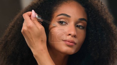 Photo for Close up female mirror reflection African American beautiful woman girl with perfect skin make-up apply oil face serum skincare product liquid cosmetic under eyes anti-aging wrinkle moisture formula - Royalty Free Image