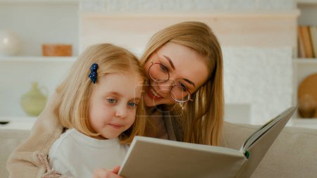 Foto de Caucasian mother in eyeglasses with little small cute daughter preschool child girl baby reading book in living room at home mom with kid read literature at couch sofa elementary learning education - Imagen libre de derechos