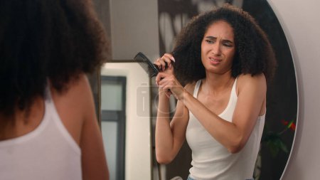 Foto de Afro americana hermosa mujer atractiva chica triste cuidado del cabello problema mirando espejo reflexión molesto mujer sufrimiento seco cabello tratando de cepillar peine enredado dañado rizos desordenados problema peinado - Imagen libre de derechos