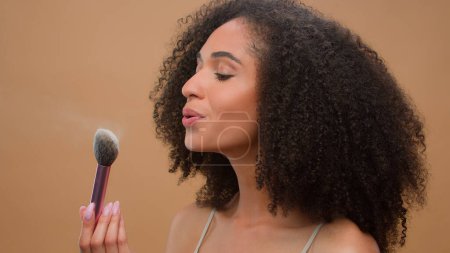 Foto de Primer plano retrato afroamericano mujer étnica modelo chica sosteniendo cepillo soplar lejos polvo aplicar el tono de piel en polvo a la nariz sonriendo a la cámara en beige estudio fondo hermosa dama cosméticos maquillaje - Imagen libre de derechos