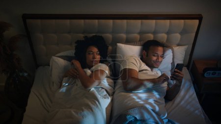Foto de Afroamericano pareja familia en cama dormitorio en la noche enojado frustrado mujer esposa tratando de dormir ofendido en marido hombre gadget adicto chatear con teléfono riendo móvil smartphone adicción problema - Imagen libre de derechos