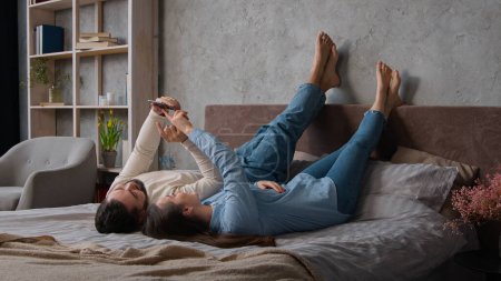 Photo for Carefree Caucasian couple in love lying down kick up feet on bed family weekend relaxing in bedroom man husband and woman wife use mobile phone application make internet order online shopping at home - Royalty Free Image