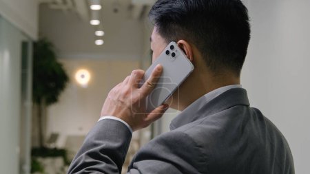 Téléchargez les photos : Arrière vue Asiatique homme d'affaires parler dans le couloir de bureau homme d'âge moyen entrepreneur professionnel chinois coréen homme parler téléphone mobile discussion d'affaires négociation tourner la tête converser dire communiquer - en image libre de droit