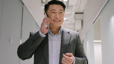 Téléchargez les photos : Asiatique homme d'affaires d'âge moyen entrepreneur adulte dans le couloir de bureau parler téléphone mobile négocier par smartphone souriant coréen japonais appelant téléphone portable heureux rire profiter de la conversation - en image libre de droit