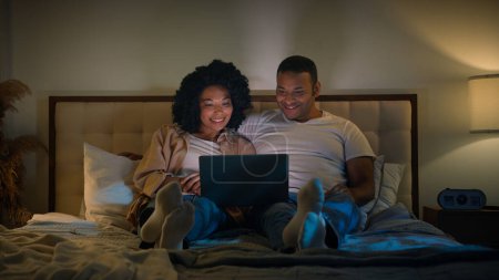Téléchargez les photos : Afro-américain couple famille homme et femme au lit nuit maison sombre chambre soir regarder portable tv film regarder film relaxant romantique date amour relation ordinateur vidéo shopping en ligne ensemble - en image libre de droit