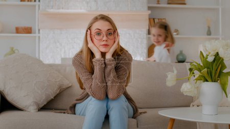Téléchargez les photos : Tired upset Caucasian mother sitting on sofa at home sad frustrated stressed exhausted fatigued mom suffer headache with active noise difficult kid child baby girl daughter running around playing - en image libre de droit