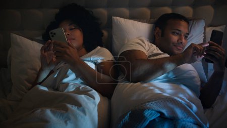 Téléchargez les photos : Gadgets addiction internet afro-américain couple homme de famille femme petite amie petit ami femme mari la nuit dans un lit confortable maison en utilisant les téléphones mobiles défilement smartphones en ignorant l'autre - en image libre de droit