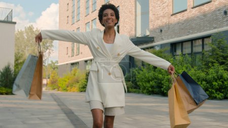 Téléchargez les photos : Afro-américaine heureuse femme marchant en ville à partir de boutique boutique boutique filature danseuse shopper fille acheteur aller client marcher à l'extérieur danse tourner avec sacs achats discount - en image libre de droit