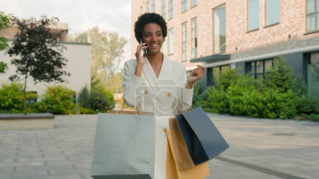 Téléchargez les photos : Afro-américain heureux souriant femme marche dans la ville magasin boutique parler téléphone mobile shopper fille acheteur femme aller client marcher à l'extérieur avec des sacs achats discount répondre appel - en image libre de droit
