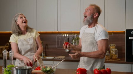 Photo for Happy Caucasian senior old couple having fun cooking at home funny man fooling around juggling tomatoes smiling woman cook fresh vegetable salad in kitchen playful family juggle natural food products - Royalty Free Image
