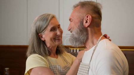 Photo for Middle aged family Caucasian gray-haired retired couple homeowners woman wife and man husband mature old grandparents cuddling hugging looking at each other at home kitchen love tender relationship - Royalty Free Image