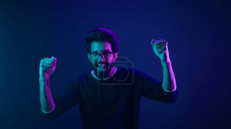Photo for Happy Indian man in ultraviolet neon studio background champion coding achievement level up celebrating winning championship fan support victory supporter cheering competition excited goal spectator - Royalty Free Image
