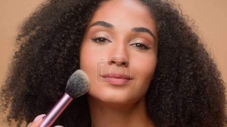 Photo for Close up portrait beige studio background beautiful African American woman with perfect smooth skin apply powder looking at camera attractive ethnic girl powdering face make-up cosmetics blush beauty - Royalty Free Image