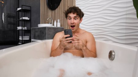 Téléchargez les photos : Homme caucasien homme nu mâle détente dans le bain avec mousse savonneuse détente repos dans la salle de bain baignoire étonné surpris surpris regarder smartphone lecture de bonnes nouvelles dans mobile wow réaction étonnante baignoire de lavage - en image libre de droit